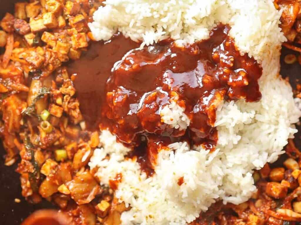 Close-up of a dish featuring white rice topped with a rich, dark sauce, accompanied by a savory mix of chopped ingredients including tofu, vegetables, and herbs, creating a vibrant blend of textures and colors.
